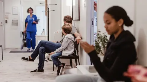 Getty Images Patients wait at a hospital as a healthcare professional walks in the corridor