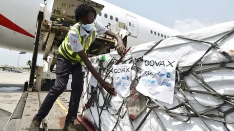 Getty Images A woman puts on Covax stickers as workers unload a shipment of AstraZeneca Covid-19 vaccines