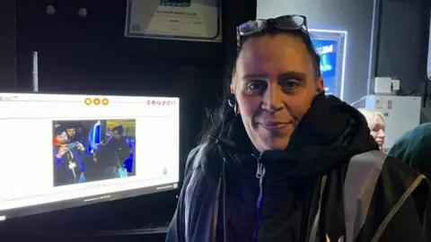 Ollie Conopo/BBC Woman with short hair and glasses on top of her head sits next to a monitor screen