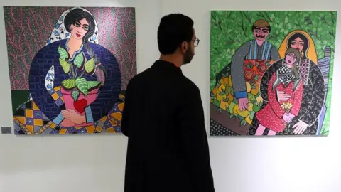Mohamed Messara/EPA A man stands between two paintings in a gallery (on the left is a painting of woman; on the right is a painting of a man, woman and child) in Tunis, Tunisia - Saturday 27 May 2023
