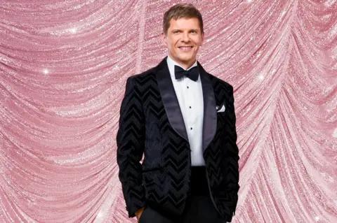 Kieron McCarron/BBC Nigel Harman in a black tuxedo with his hands in his pockets