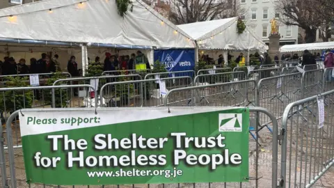 BBC Sign saying "The Shelter Trust for homeless people" on railing in front of marquee, people inside buying soup