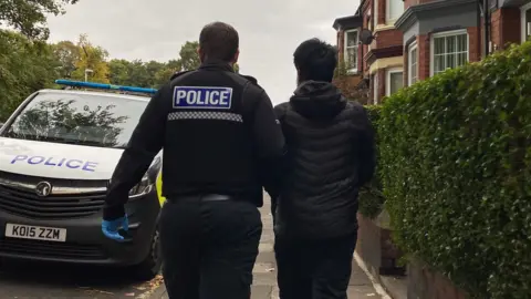 BBC A suspect in black clothes is led to a police van by an officer