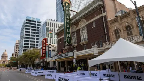 Getty Images A brick building on a long street with security personnel outside and white barriers in front with SXSW printed on them in black. A large, green vertical sign with the word "Paramount" is attached to the front of the building. In the distance we can see orange and white barriers, showing that the road has been blocked off to allow pedestrians to move around freely.