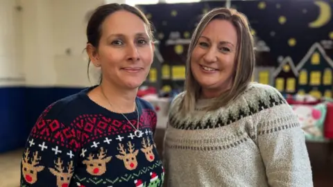 Laura Haynes and Jess Draper. Laura has dark hair and is wearing a navy Christmas jumper printed with red and white patterns and reindeer heads. Jess is blonde and is wearing a cream jumper with a pattern across the shoulders. Both women are smiling at the camera.