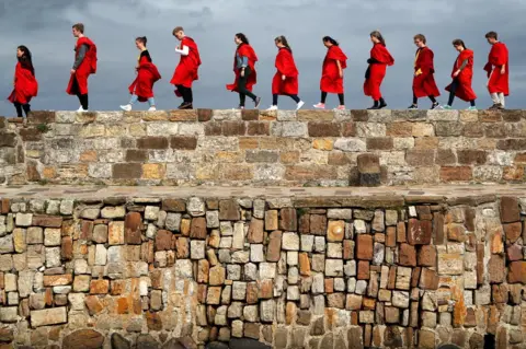 Jane Barlow/ PA A row of students in red gowns walk along the harbour wall.