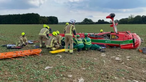 A red light aircraft upside down. Wheels are sticking up and it has been surrounded by emergency services including paramedics and firefighters