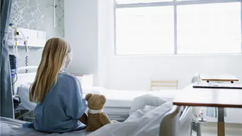 Getty Images Young girl sitting on a hospital bed with a teddy bear next to her