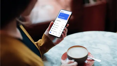 Getty Images Woman on online banking app on her smartphone paying for a coffee in a cafe