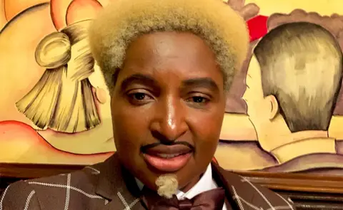 Don One A selfie of Don One, who has golden hair, wearing a checked brown suit. he is looking into the camera with the background of a painting of two people looking at each other