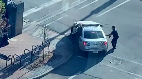 A police officer with his gun drawn takes cover behind a police car