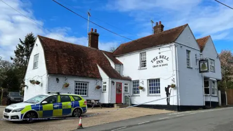 The Bell Inn in Feering, Colchester. The building is white, and there is an Essex Police car in front of the property and a police cordon has been put in place