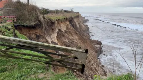 ANDREW TURNER/BBC A farm gate hangs over the edge of the crumbling cliff at Happisburgh