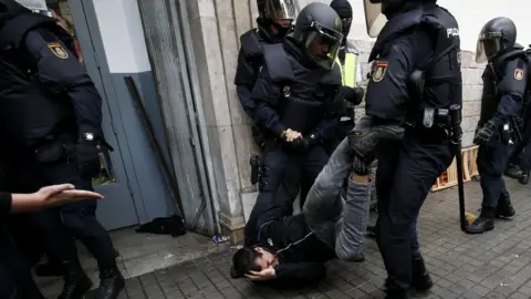 Getty Images A man, lying on the ground, clutches his head in pain as he is dragged through the street by his legs by two police officers in full riot gear