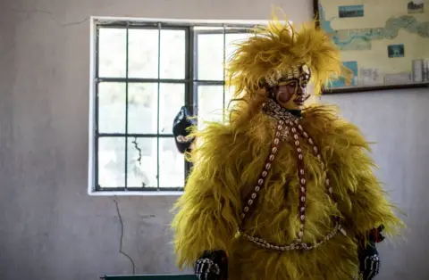 AFP Man wearing the Senegalese Simba mask. He is wearing a bright yellow furry outfit, and has a painted face