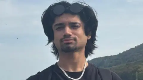 Supplied Aryan Sharma, 20, with collar-length black hair. He's wearing black round shades on his head and a sliver chain with a clear sky behind him.