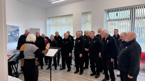 A mens choir, all stood in a semi-circle in black zip up jumpers and black trousers. The conductor is facing away from the camera with a music stand in front of her. She has short blond hair and is wearing black trousers and a grey and orange jumper. The flood is wood, there is a keyboard in the left bottom corner. There are large windows in the background with curtains.