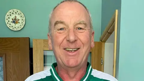 Paul Tudor A man smiling at the camera while wearing a green Wolverhampton Casuals FC shirt featuring the club badge, a 1899-2024 anniversary crest and the sponsor Vogue Ceramics. He looks to be stood in a kitchen. 