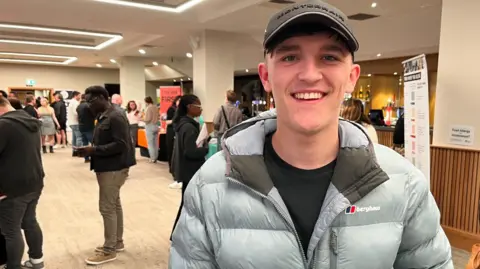  Alfie, 17, at a jobs fair in Sunderland. He smiles at the camera and wears a grey Berghaus puffer jacket and peaked cap.