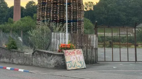 A bonfire of wooden pallets sits behind a fence, in front of the fence is a brick wall and a wooden sign is propped up against it with "bonfire 10th July" written on it