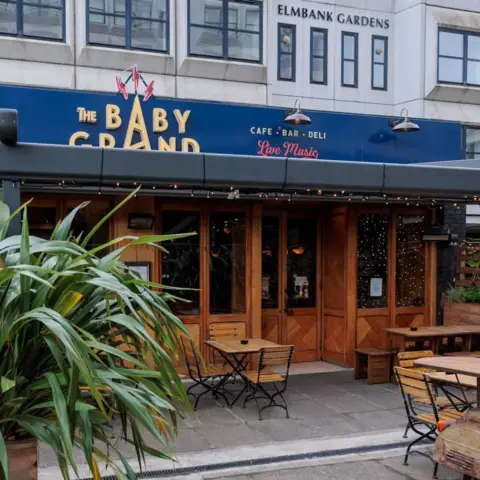 The Baby Grand An outside view of the Baby Grand piano bar. There is a plant in the foreground. There are tables and chairs outside. A sign above the door, which is surrounded by wood, is blue, with the name of the bar in yellow. Another sign, reading 'Elmbank Gardens' is up high in the background on a light-coloured building.