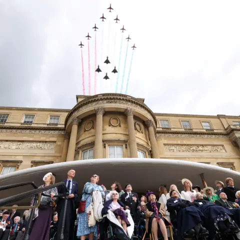 Reuters Red Arrows deploy coloured smoke as they fly over Buckingham Palace with a group of veterans all looking up to the sky below. It's a moody, grey cloudy day.