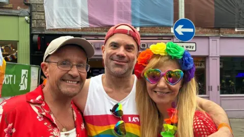 Three people smile, with their arms around each other. They wear bright rainbow-coloured clothing and accessories.