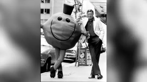 A black and white picture of Roger Hargreaves leaning on a person in a Mr Man costume