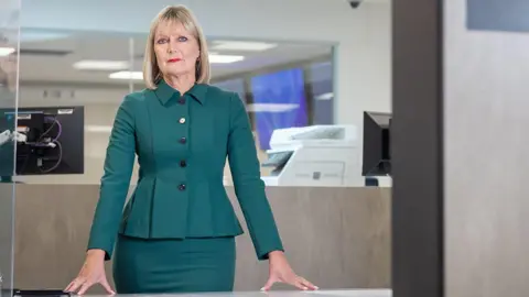 Joy Allen staring into the camera with her hands resting on a surface in front of her. She's got shoulder-length blonde hair and is wearing bright red lipstick. She's wearing a dark jacket that some people would say is blue and others green, with a matching skirt. She's standing in what looks like a police station. There are computers behind.