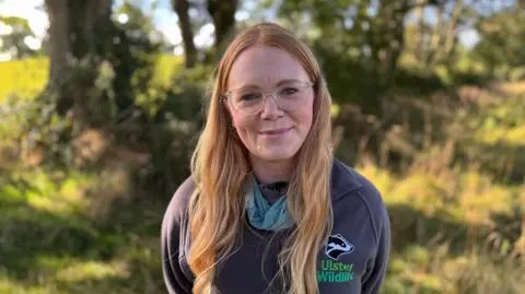Katy Bell has long strawberry blond coloured hair and is wearing glasses and an Ulster Wildlife fleece. She is standing in front of long grass and trees