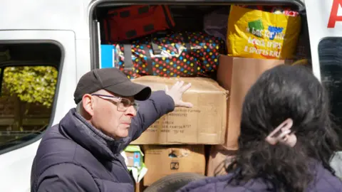 Diocese of Northampton A man in a black puffer jacket and black cap raises his arm towards lots of cardboard boxes stacked up in an ambulance. 