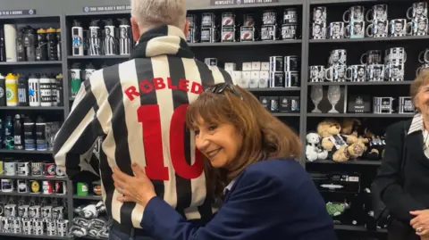 Elizabeth Robledo hugging a man who is facing away from her so only his back is visible. He is wearing a black-and-white football shirt with "Robledo, 10" on the back. Behind them are shelves stacked with football memorabilia.