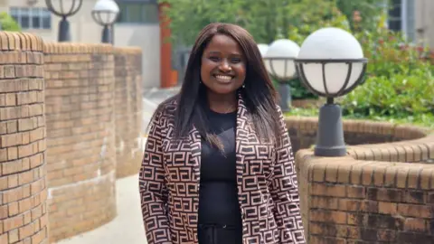 Louise is smiling at the camera. She has dark brown hair and is wearing a jacket with a black and orange square-like pattern.