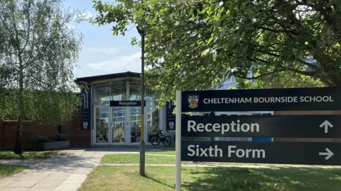 The front of Bournside School in Cheltenham. In the foreground of the image is a sign which shows the school name and crest with 'reception' and an arrow point upwards, and below 'Sixth Form' with an arrow to the right. Bind you can see the glass front of the school with a 'reception' sign above. There is a path up to the entrance of the school with grass either side.