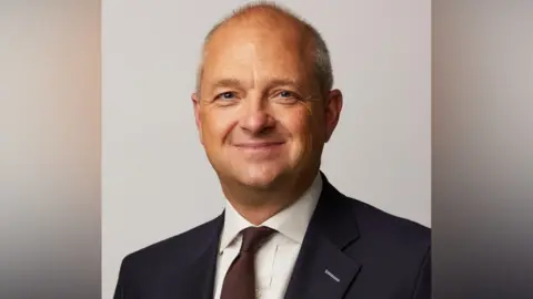 UK Parliament Jerome Mayhew looks towards the camera and smiles, wearing a dark suit, white shirt and tie