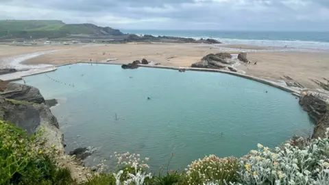 An aerial shot of Bude Sea Pool with frilly sea vegetation in the foreground. The water is clear and blue-green and there is a beach and sea behind it.