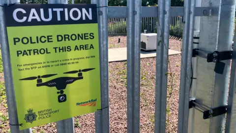 A sign is attached to a metal fence. It reads 'Caution. Police drones patrol this area." In the background, there is a white box containing the drone on an area of gravel.