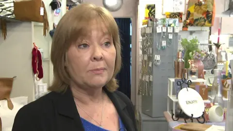 Janene Stavert, manager at La Maison Boutique in Oxted, being interviewed in her shop. She is wearing a black suit jacket and a blue top. Some of the shop's stock can be seen behind her, including jewelry, picture frames and bags.