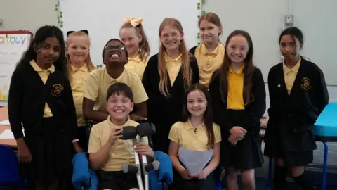 Shaun Whitmore/BBC A group of ten primary school children having their photograph taken. There are eight children across the back row and two children sitting down at the front. They are wearing their school uniform which consists of yellow polo shirts. They are standing inside a classroom.