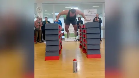 A man in gym clothes pictured mid-air between two towers of foam blocks about a metre high.