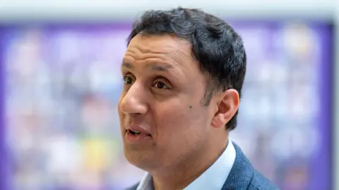 PA Media Anas Sarwar speaking at an event- he has dark black hair and is wearing a suit.