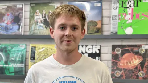 Raphael Sheridan / BBC James, 25, with blonde hair wearing a cream T-shirt is smiling at the camera in front of row of records