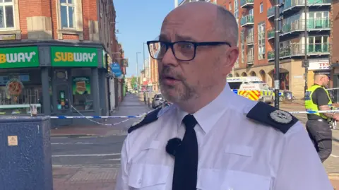 Oli Constable/BBC A police officer wearing a white shirt with a black tie and lapels. He is standing in front of a crime scene.