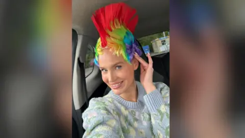 Sophie Jones A woman has a rainbow mohawk wig on. She has her hand on her head and she is smiling at the camera. She is wearing a blue jumper on with flowers.