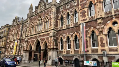 The exterior of the Royal Albert Memorial Museum in Exeter, which is in the style of Victorian Gothic Revival. The museum's exterior uses sandstone, has detailed stonework and uses characteristic Victorian features, including arched windows and ornate gables. People are walking past