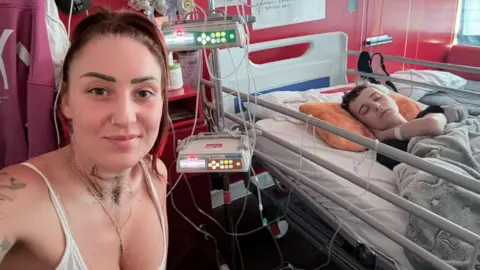 Family A woman with brown-red hair tied back and wearing a white strappy top sits next to a hospital bed where the boy from the above photos is lying hooked up to machines.