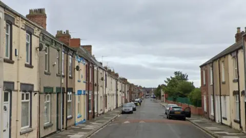 A residential street of terraced houses with cars parked along the sides.