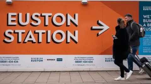 PA Media Passengers with suitcases walk past a billboard