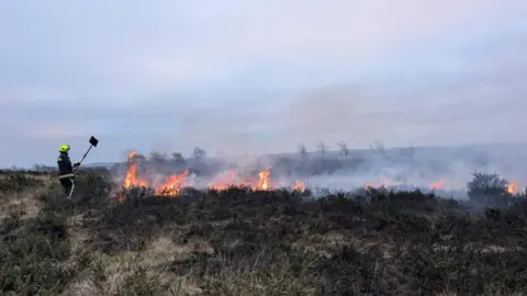 Devon and Somerset Fire and Rescue Service A firefighter stood to the left of a large fire on the moors. In the distance are bright orange flames with a large amount of smoke. The moorland is black with ash. 