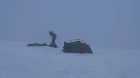 People setting up camp atop of Helvellyn in full white-out conditions. There are two people crouched trying to set up a tent.